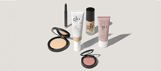 Assorted glo cosmetic products: foundation and primer tubes, a pencil, and two powder compacts on a light background.