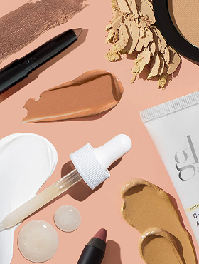 Makeup flatlay with cream and powder swatches, dropper, compact crumbs, lipstick pencil on peach background; tube labeled 'glo'
